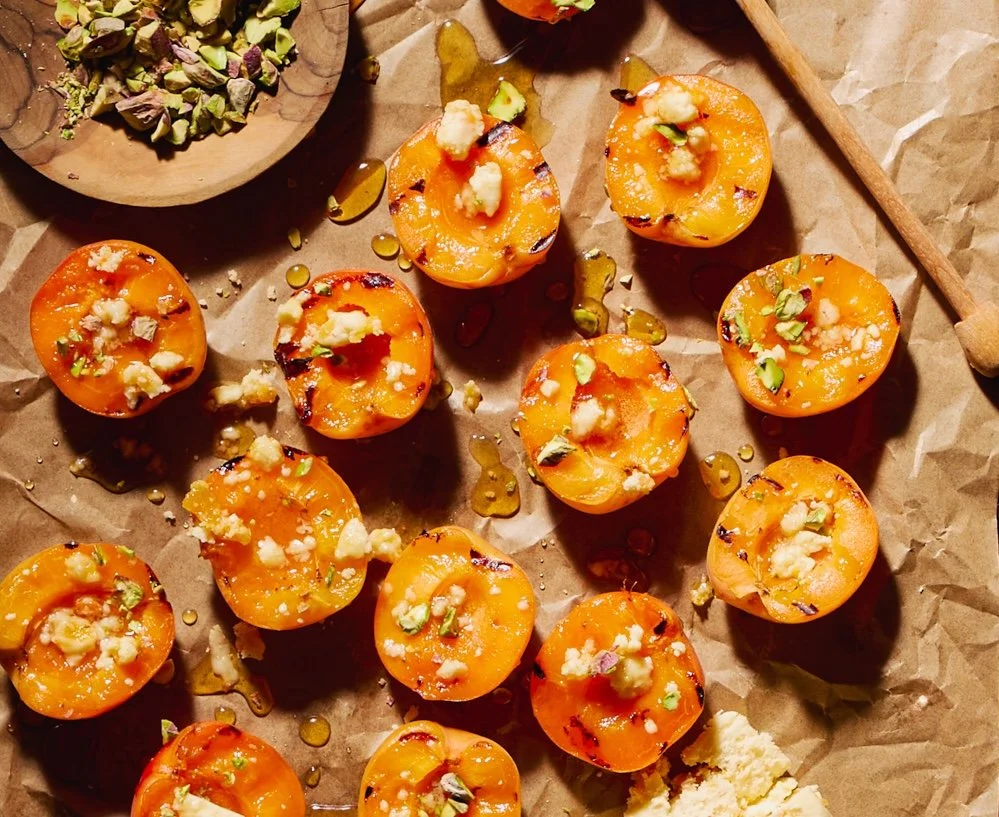 Halved grilled apricots topped with crumbled cheese, chopped pistachios, and drizzled with honey, arranged on parchment paper with a bowl of chopped pistachios and a wooden spoon nearby.