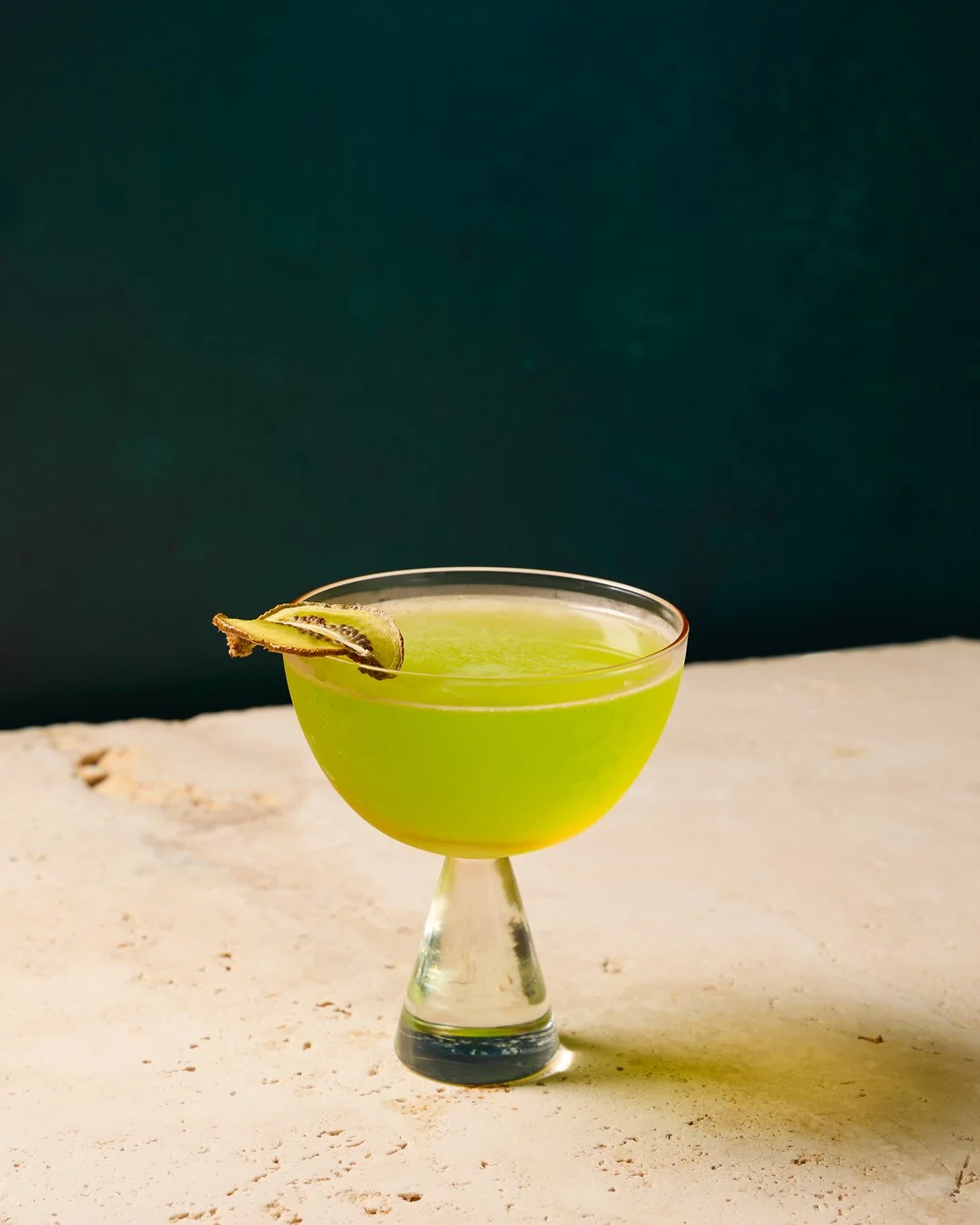 A green cocktail in a coupe glass with a garnish of kiwi slices, set on a white surface with a dark background.