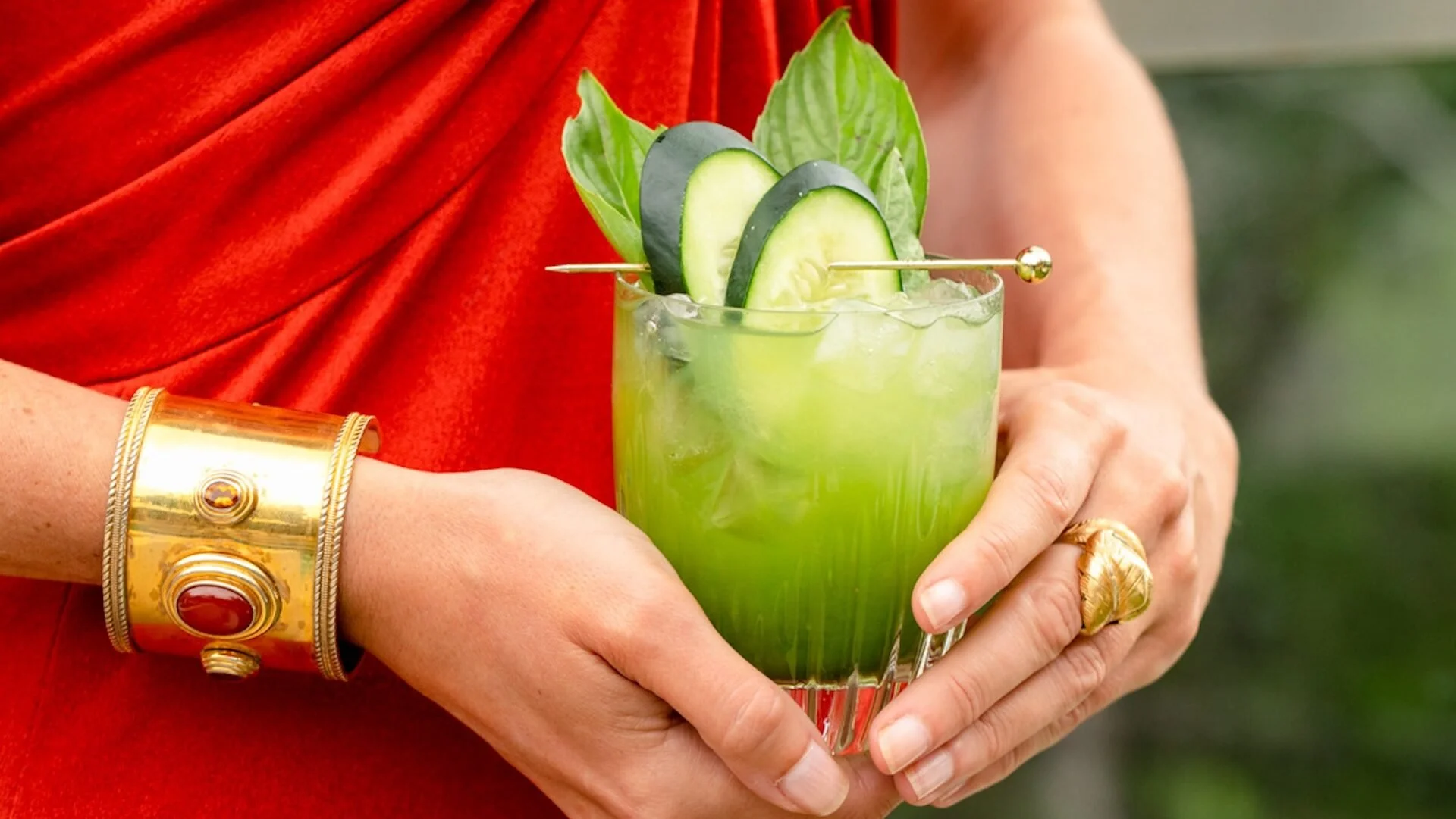 Person holding a tall glass of green cucumber and lime cocktail garnished with cucumber slices and green leaves, wearing a gold bracelet and gold ring.