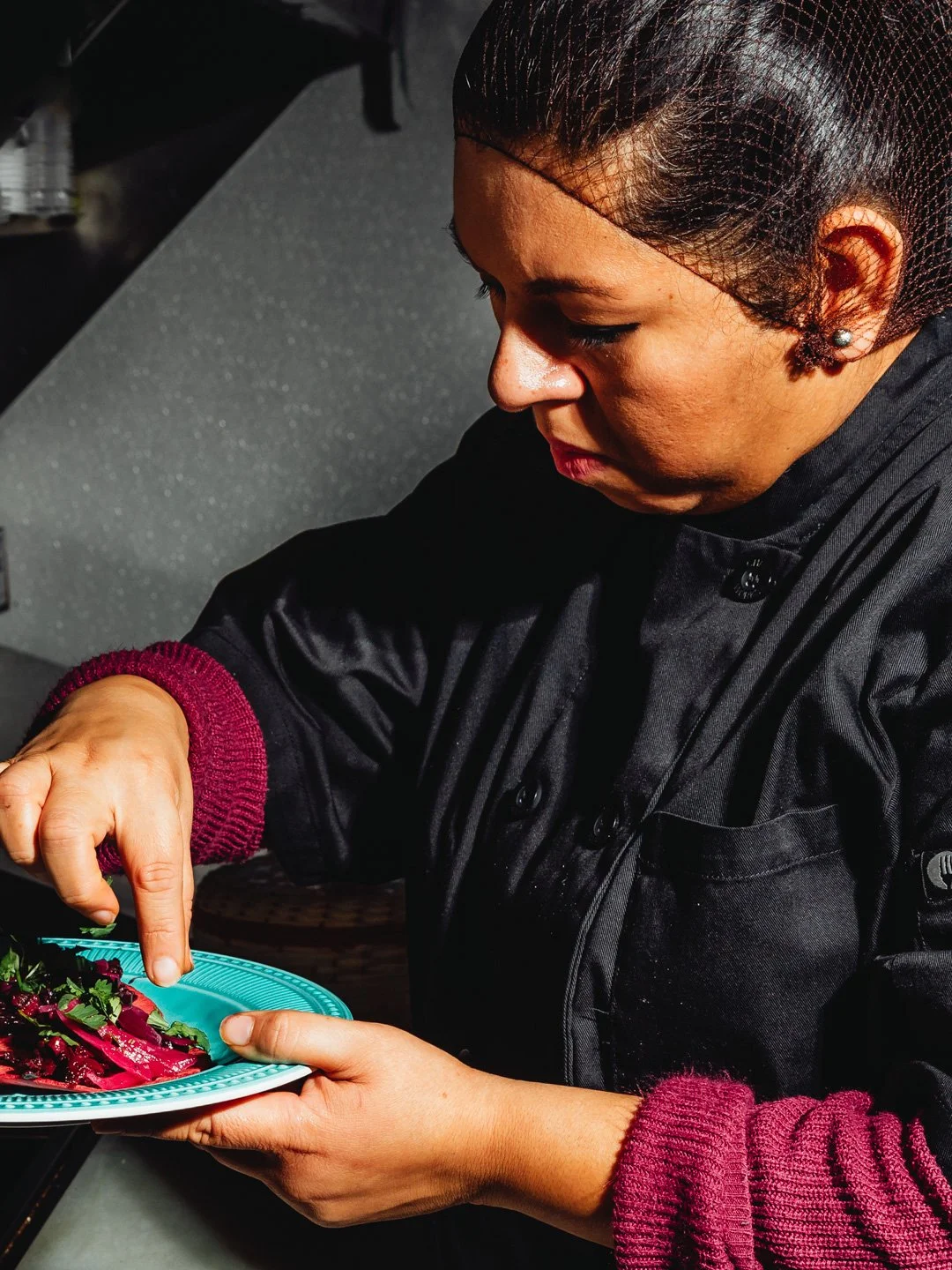 A woman in black chef's coat garnishing a plate of food, possibly in a restaurant kitchen.