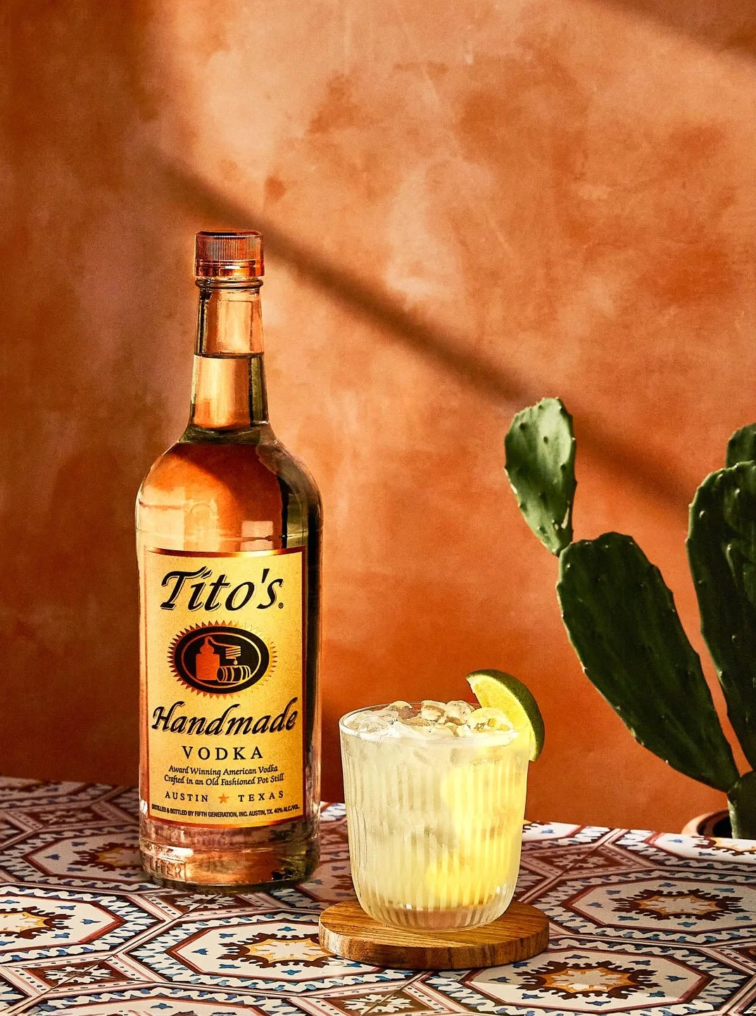 A bottle of Tito's handmade vodka, a glass of a cocktail with ice and a lime wedge, a cactus plant, and a patterned tablecloth with a warm, peach-colored background.