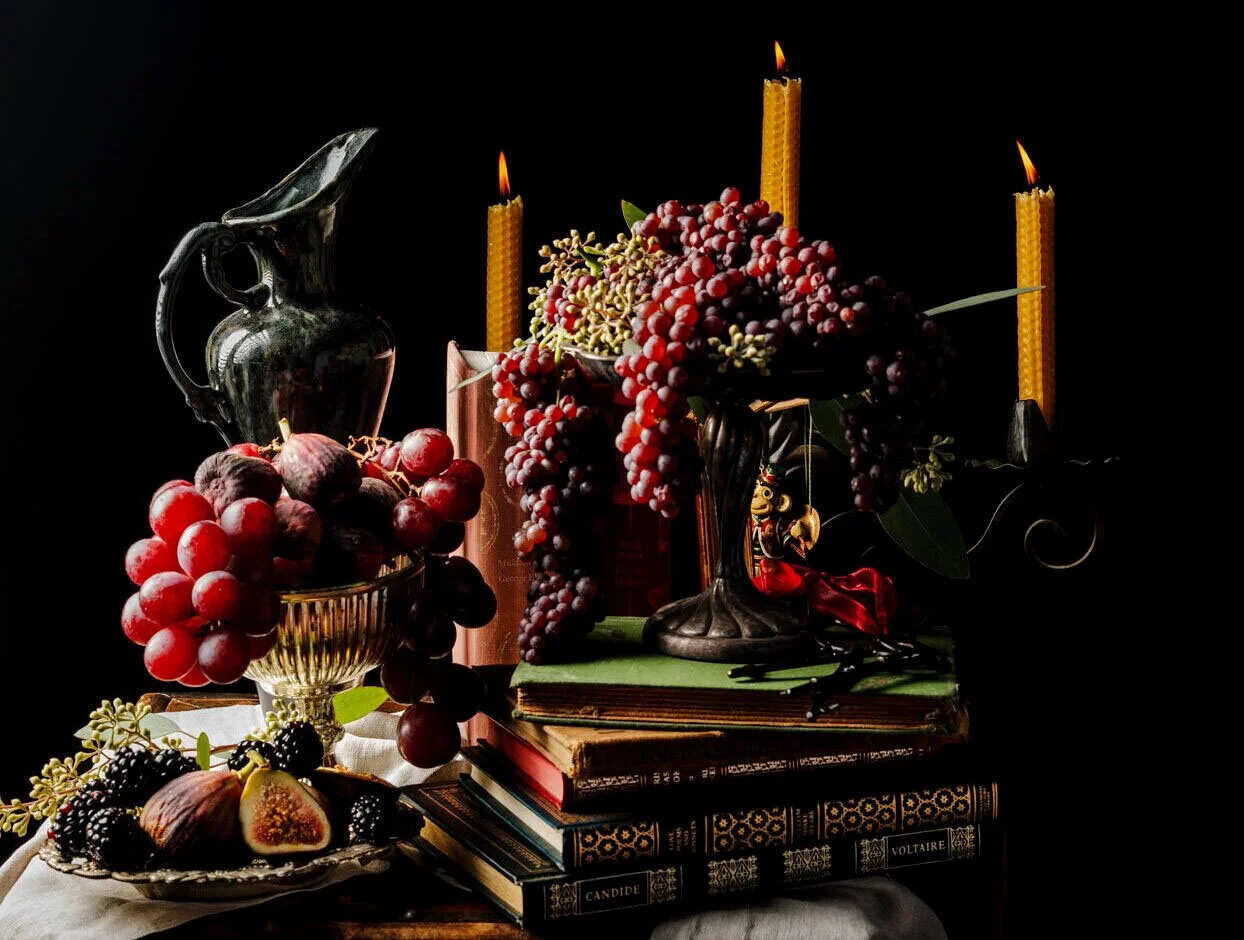Still life of grapes, figs, and flowers with books, a ceramic pitcher, and lit beeswax candles on a dark background
