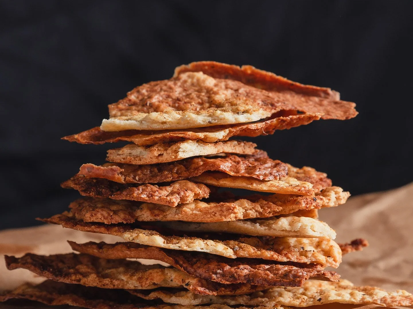 Stack of various types of crispy, cooked flatbread or crackers on a piece of parchment paper against a dark background.