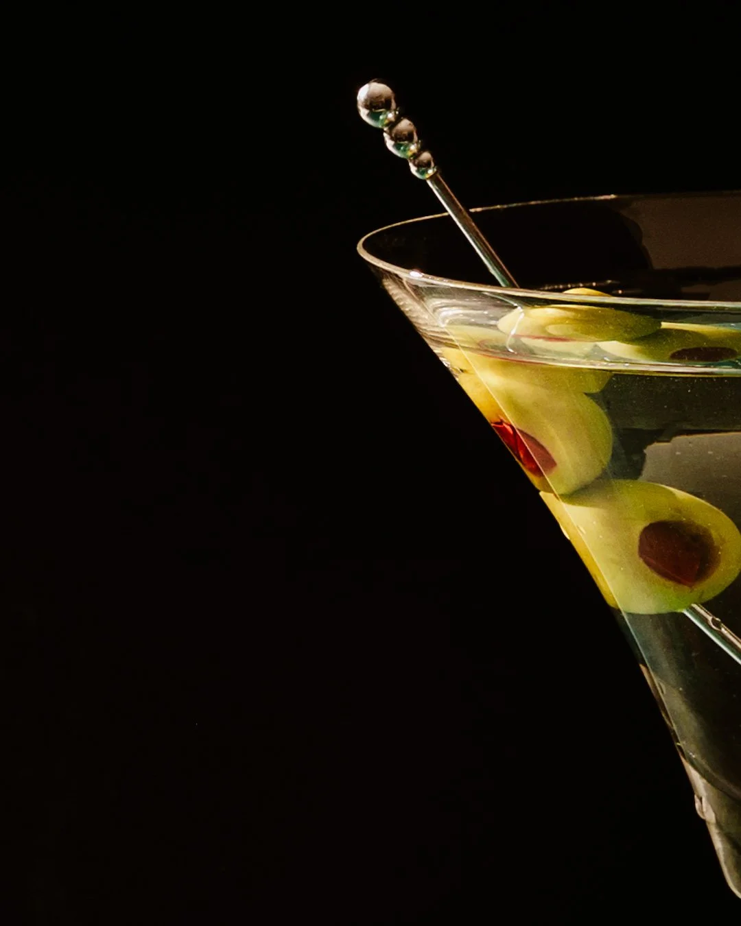 Close-up of a martini glass with green olives and a cocktail pick, against a black background.