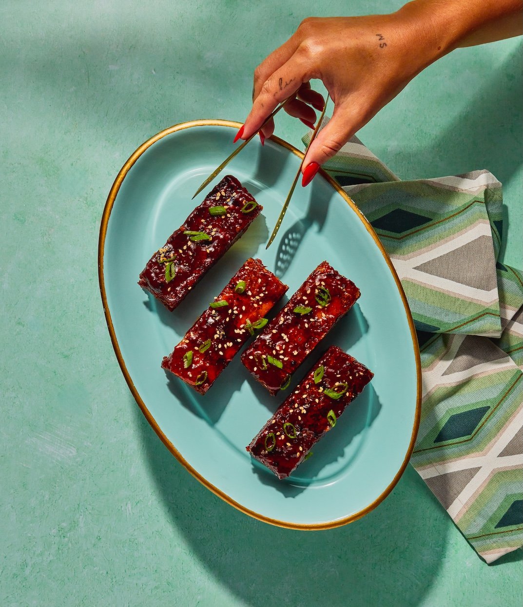 A hand with red nails holding chopsticks, picking up pieces of glazed, garnished ribs on a blue oval plate.