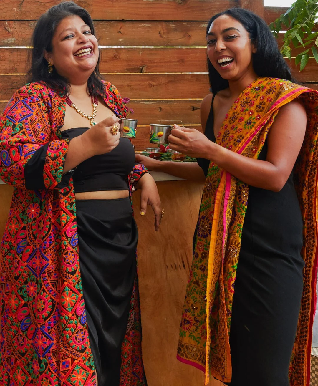 Two women dressed in colorful, traditional Indian sarees and jewelry, smiling and holding cups while standing against a wooden fence and greenery.