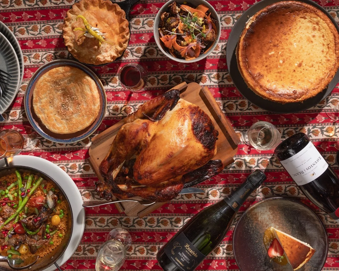 A festive dinner table with a roasted turkey, various pies including pumpkin and apple, a salad, a casserole, a bottle of red wine, and glasses of wine, all placed on a red and white patterned tablecloth.