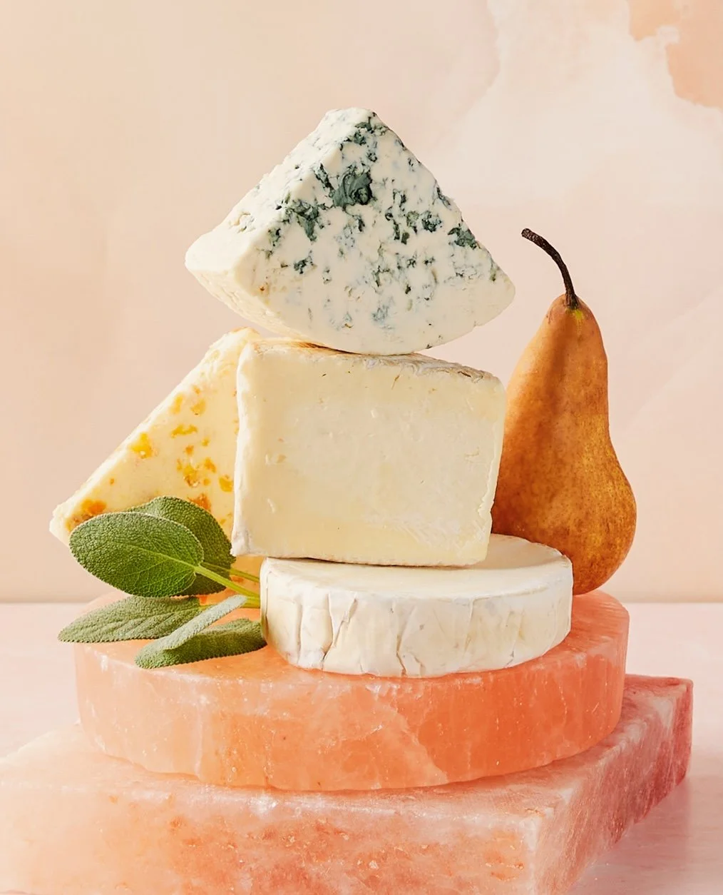 A stack of various cheeses, a pear, and sage leaves on a pink Himalayan salt block.
