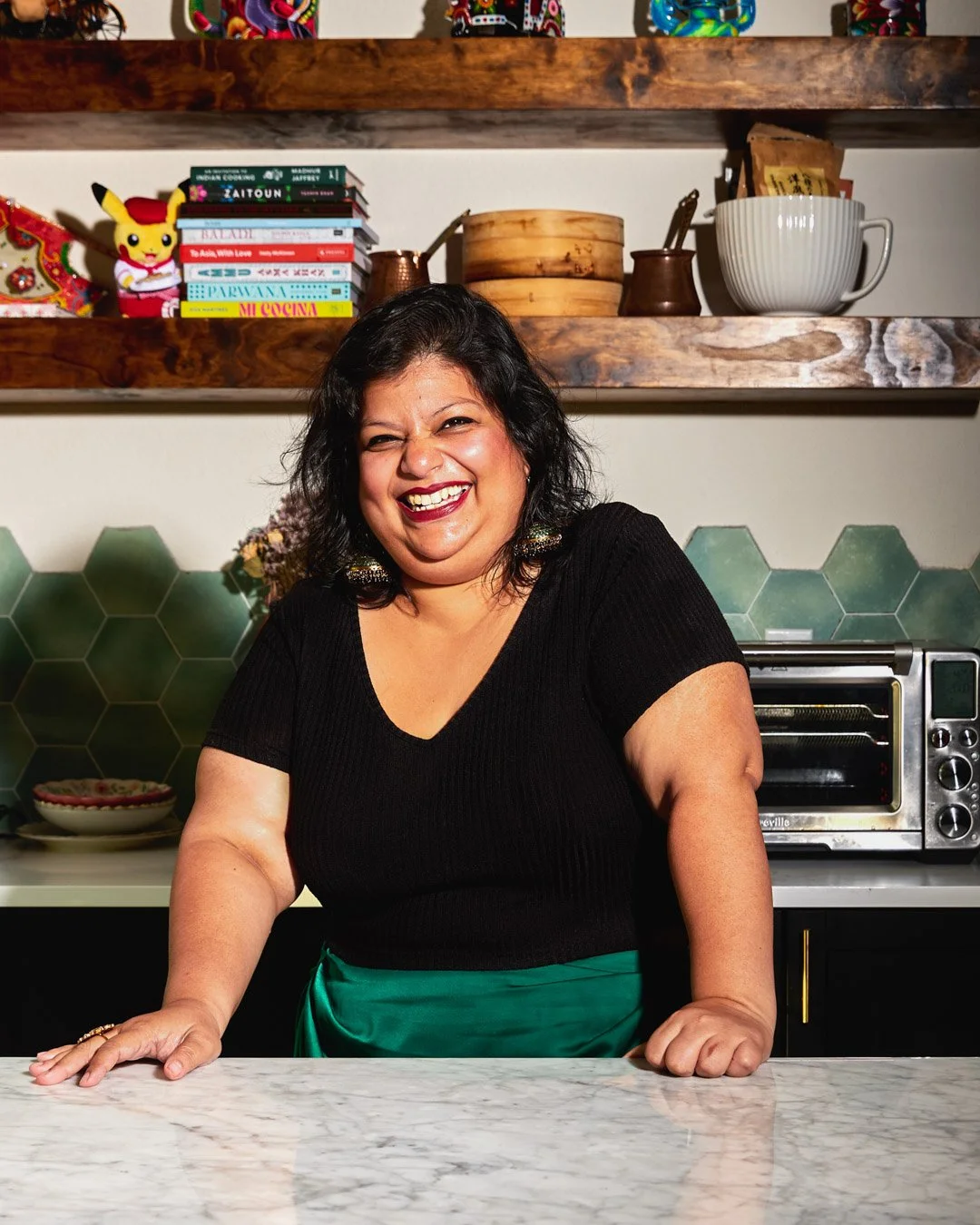 A woman with dark hair smiling and leaning on a white marble kitchen counter, wearing a black top and a green apron, with a colorful backsplash and shelves with books, decorative items, and kitchenware in the background.