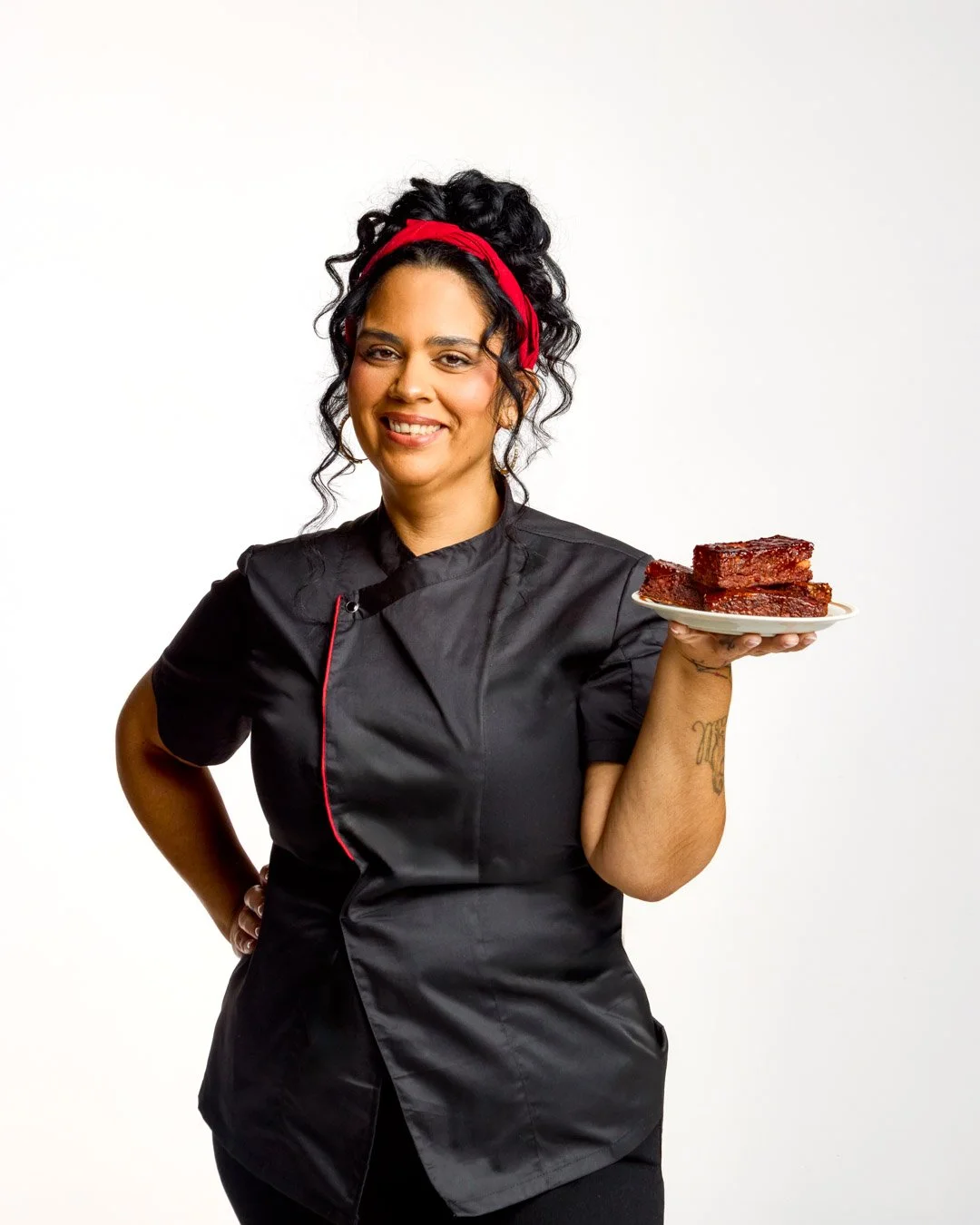 A woman dressed as a chef, holding a plate with three brownies, smiling at the camera.