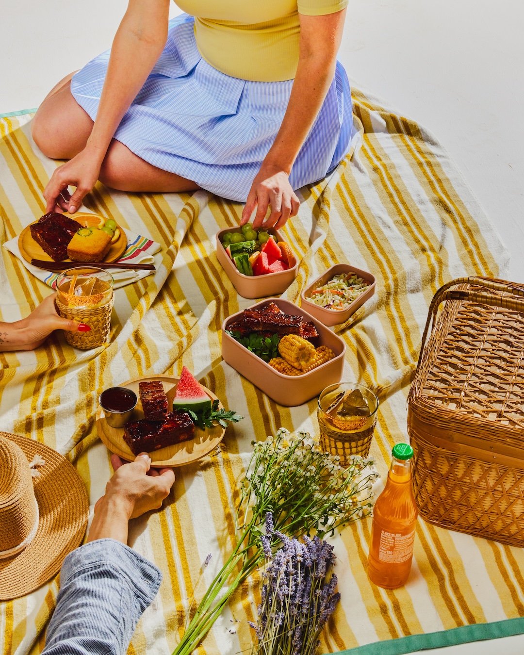 A picnic scene with a person sitting on a yellow striped blanket, surrounded by various food items, drinks, and flowers.