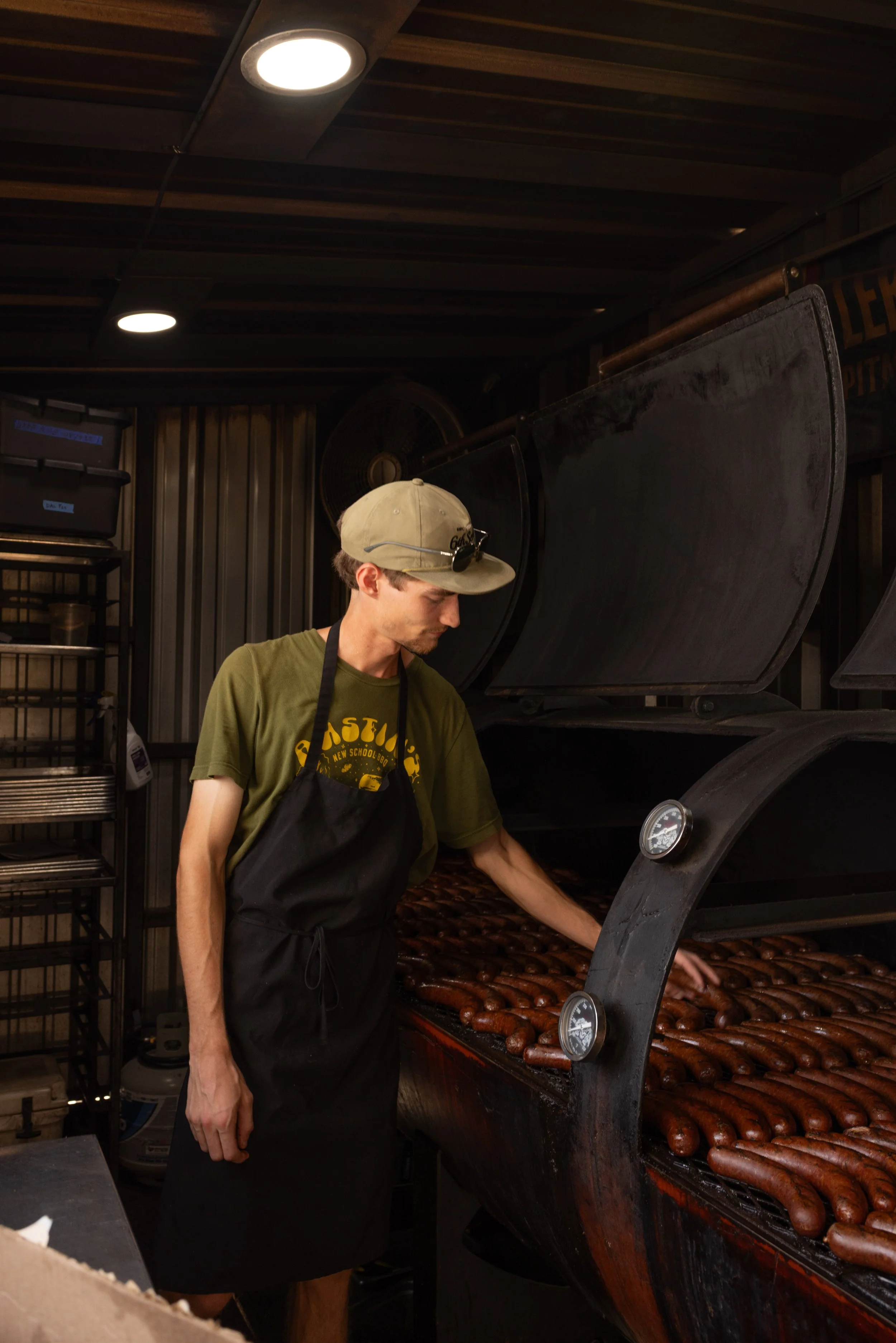 A man wearing a beige cap with sunglasses on it and a green T-shirt working at a hot dog stand or food truck. He is cooking or grilling hot dogs on an outdoor grill. The background includes a metal storage rack with trays and a large fan on the wall,