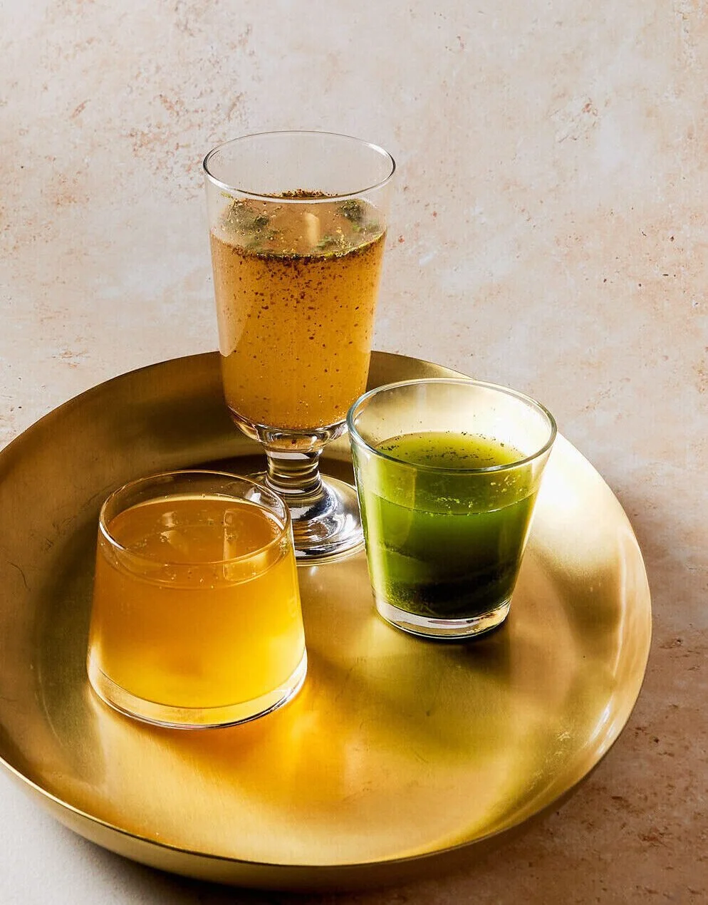 Three beverages, one in a tall glass with orange liquid and bubbles, one in a short glass with yellow liquid, and one in a short glass with green liquid, arranged on a round gold tray.