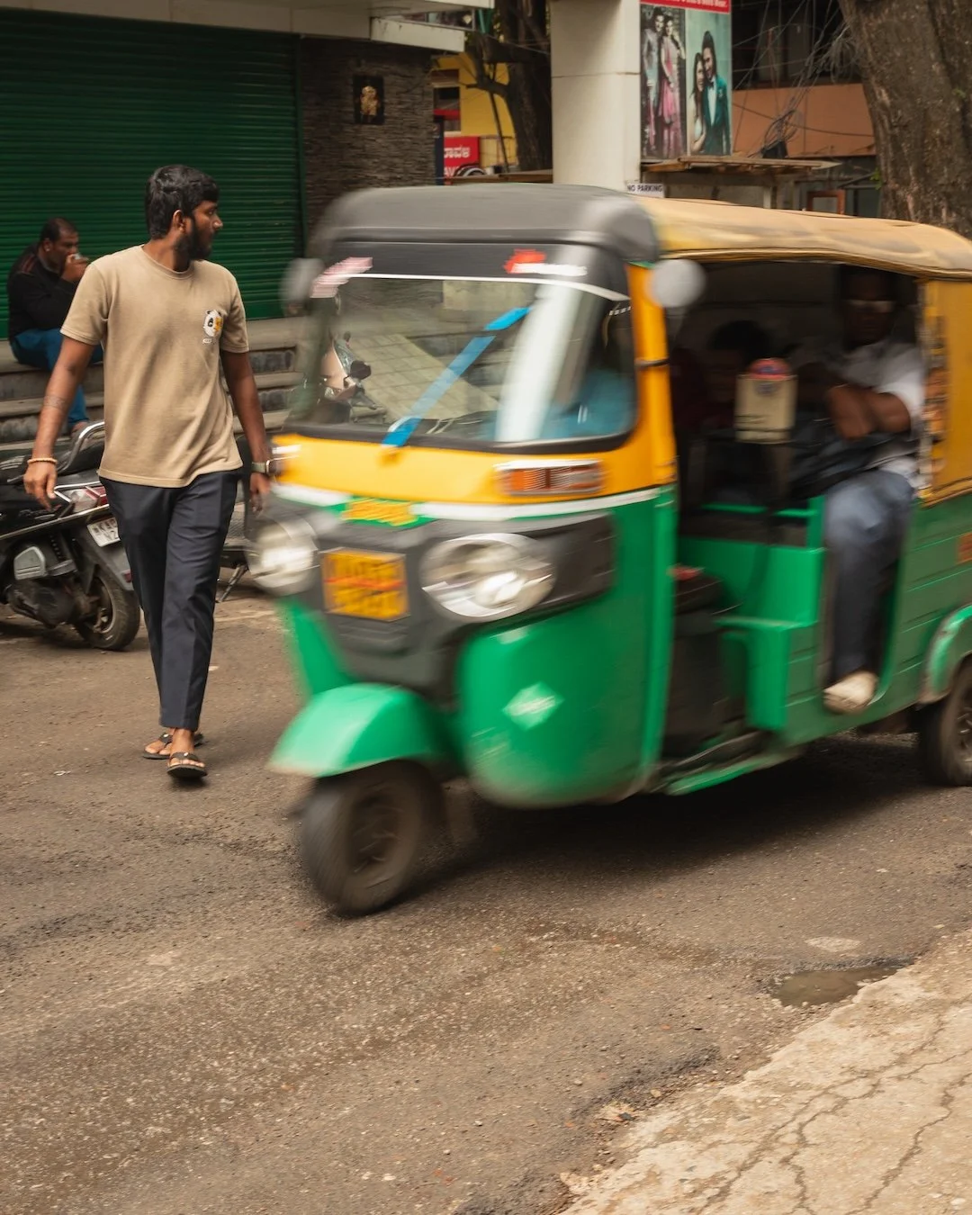 A man walks past a green and yellow auto rickshaw on a busy street, with a motorcycle and storefronts in the background.