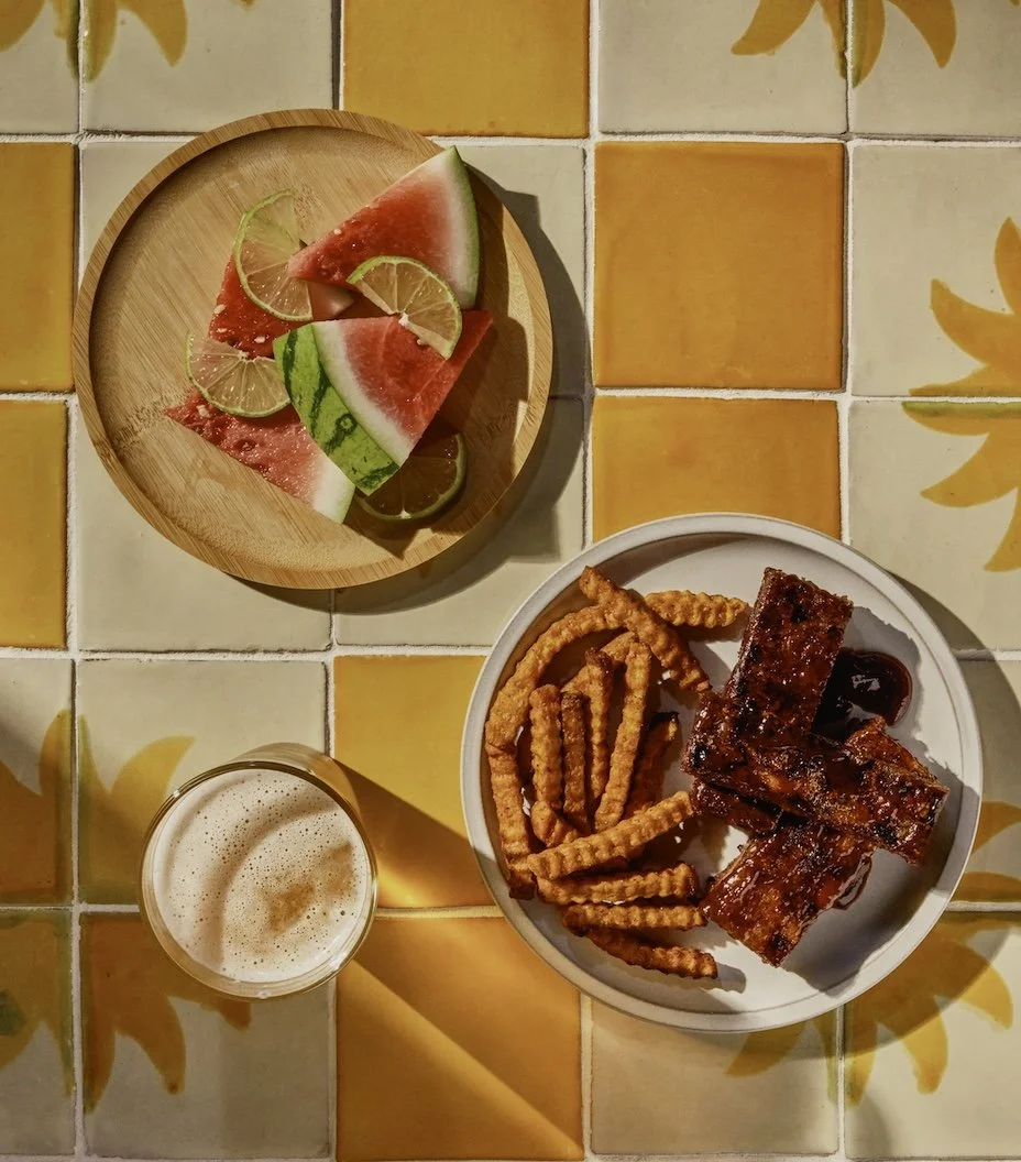 Watermelon slices with lime on a wooden plate, a glass of beer with foam, and a plate with a rack of ribs and French fries, on a colorful tiled table.