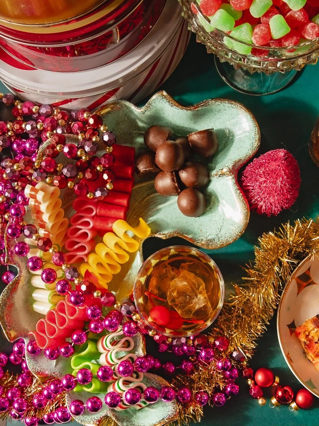 Holiday table with candy canes, chocolates, colorful beads, glass bowl with gummy candies, and a drink topped with ice, all decorated with gold tinsel and red and green accents.