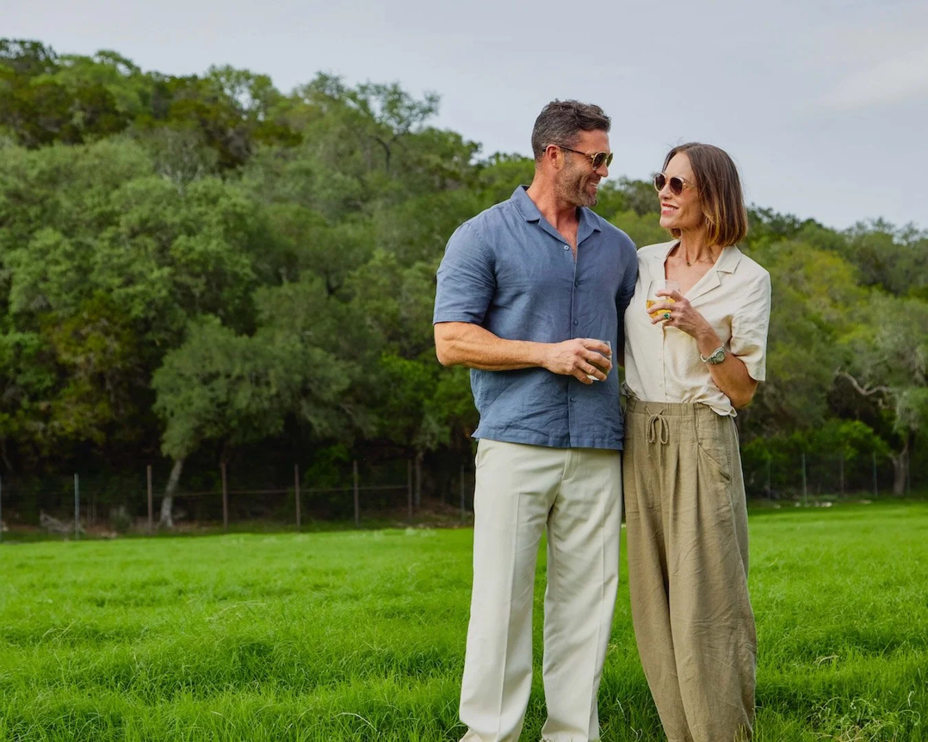 A middle-aged couple stands together on a grassy field, smiling at each other while holding drinks, with trees in the background. Lifestyle photography by Nitya Jain for San Antonio Tourism.