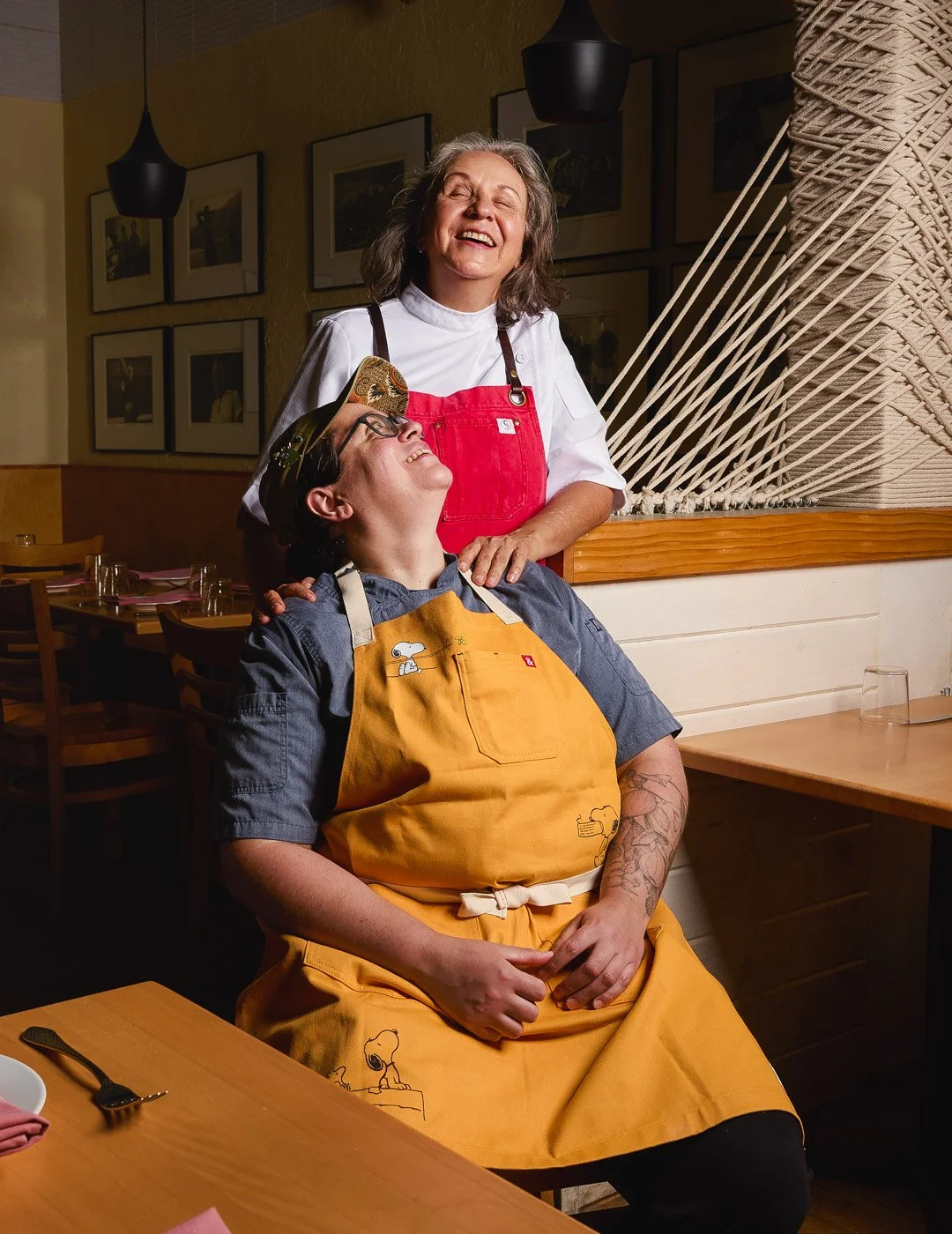 A woman in a white shirt and red apron is standing behind a man in a gray shirt and orange apron, who is sitting at a restaurant table and smiling. Both are laughing or smiling happily, with the woman resting her hand on the man's shoulder.