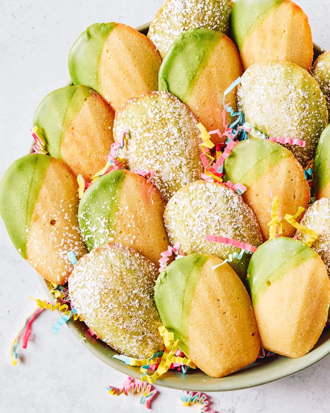 Colorful cookies, some topped with powdered sugar, decorated with green and orange icing, surrounded by colorful confetti and shredded paper in a bowl.