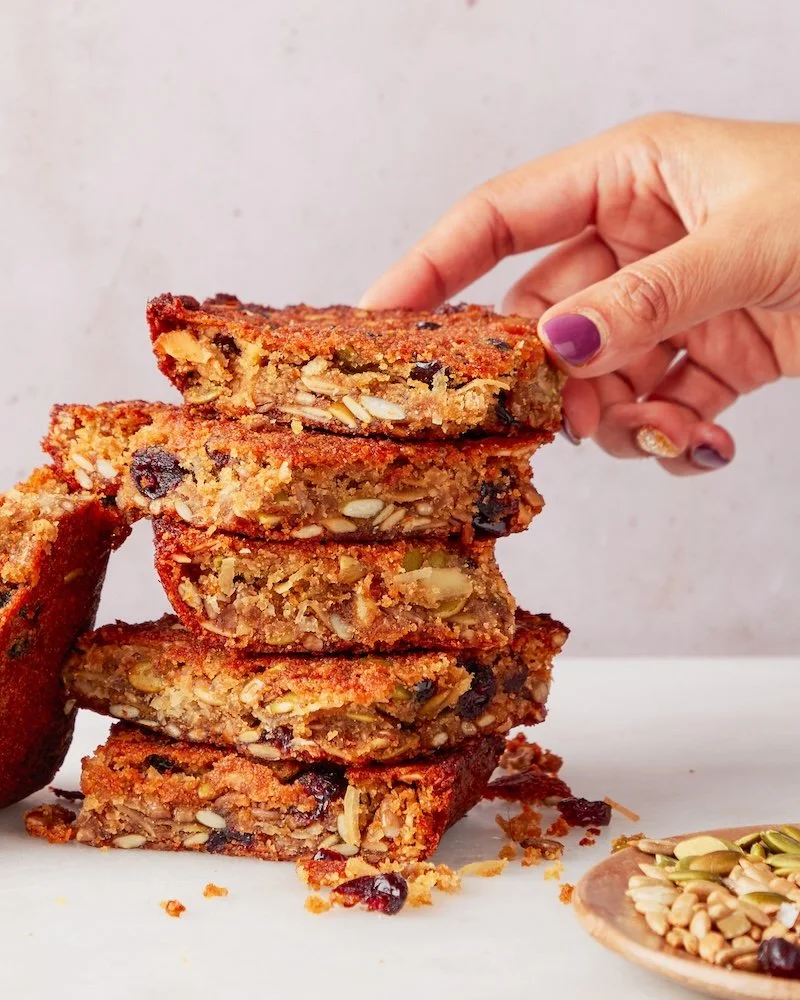 A hand stacking five slices of homemade fruit and nut bars on a white surface. The bars contain oats, seeds, dried fruit, and nuts.