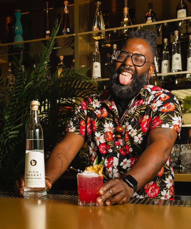 A bartender with glasses, a ring, a tattoo, and a watch is smiling and sticking out his tongue behind a bar counter. He is wearing a colorful Hawaiian shirt with a floral pattern. In front of him on the bar are a red cocktail garnished with a pineapp
