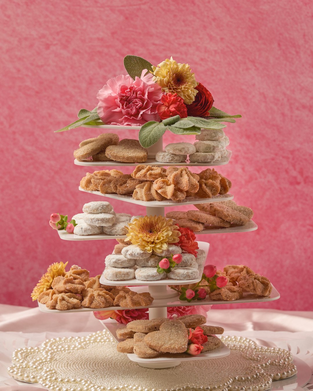 Tiered platter of assorted cookies decorated with pink and yellow flowers, pink berries, and greenery against a pink background.