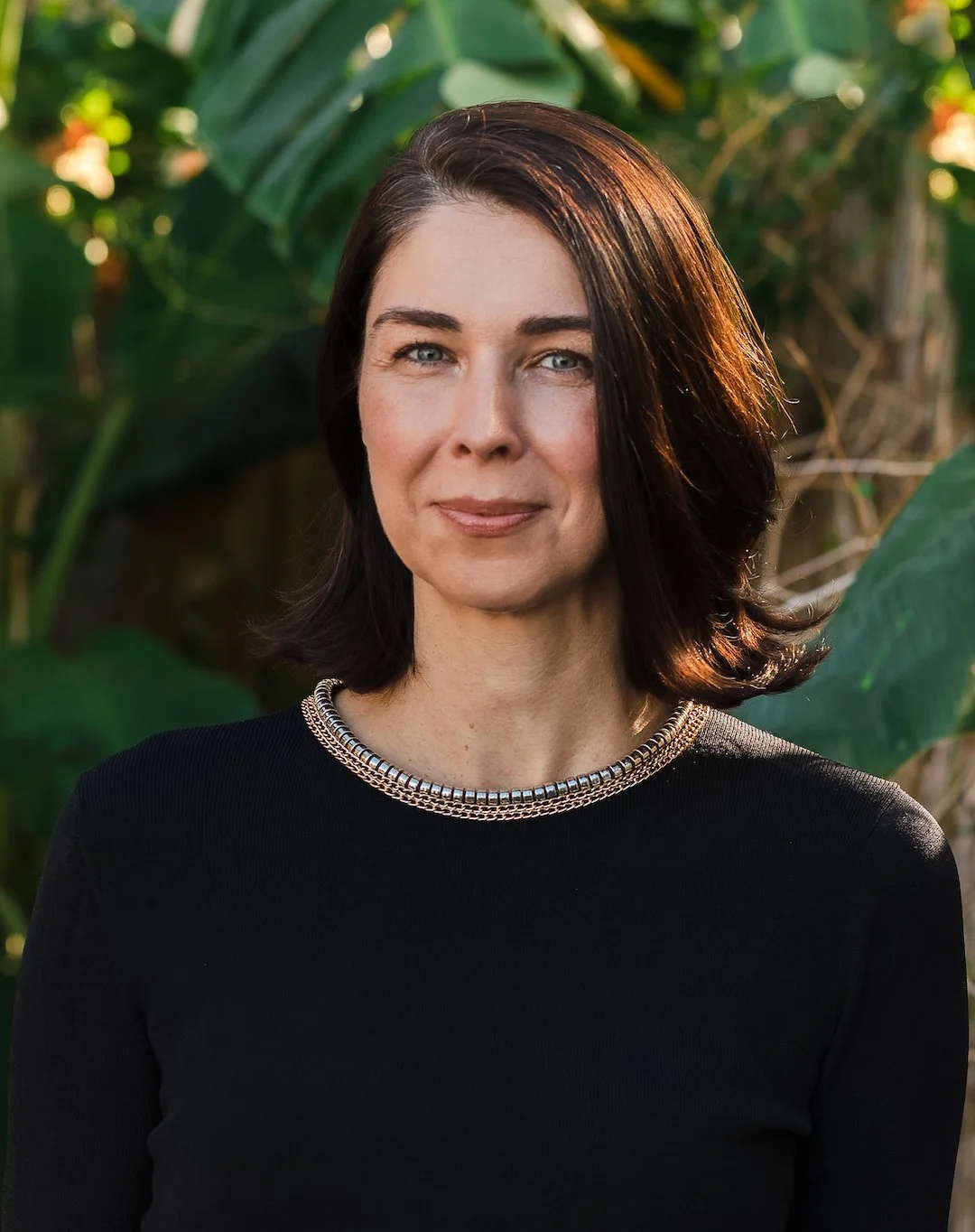 A woman with shoulder-length dark brown hair, wearing a black top with a silver necklace, stands outdoors in front of green foliage.