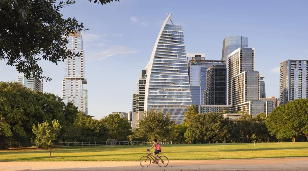 A woman riding a bicycle in a park with tall modern city buildings in the background on a clear day.