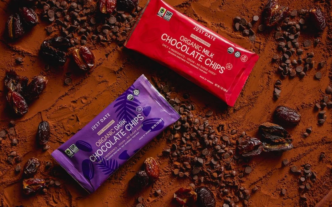 Two packages of organic chocolate chips, one dark and one milk, on a surface sprinkled with cocoa powder and scattered with dates and chocolate chips.