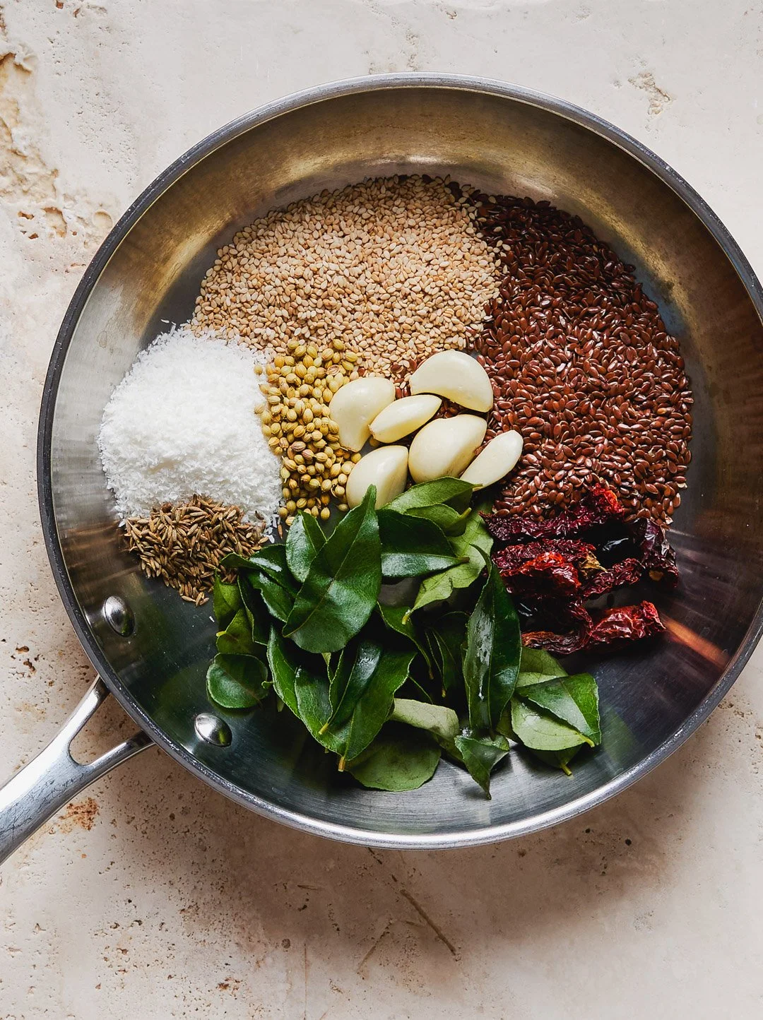 Assorted ingredients for cooking in a stainless steel pan, including sesame seeds, flax seeds, shredded coconut, coriander seeds, garlic cloves, bay leaves, sun-dried tomatoes, and dried red chili peppers.