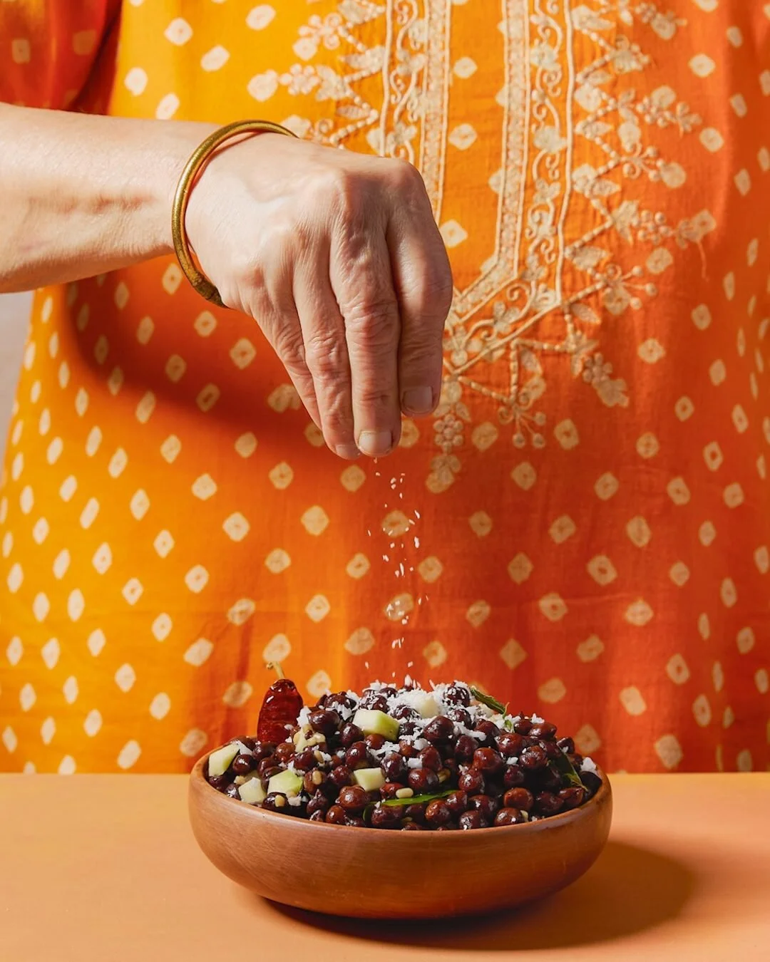 A hand sprinkling salt on a bowl of chickpeas and chopped vegetables, with the person wearing a gold bracelet and an orange, embroidered traditional garment in the background.