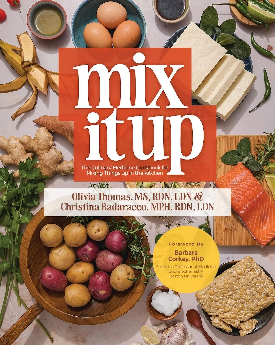 Cover of a cookbook titled 'Mix & Up' featuring an arrangement of various fresh ingredients including eggs, ginger, radishes, tofu, salmon, garlic, cilantro, microgreens, green chili peppers, and rice cakes on a kitchen counter.