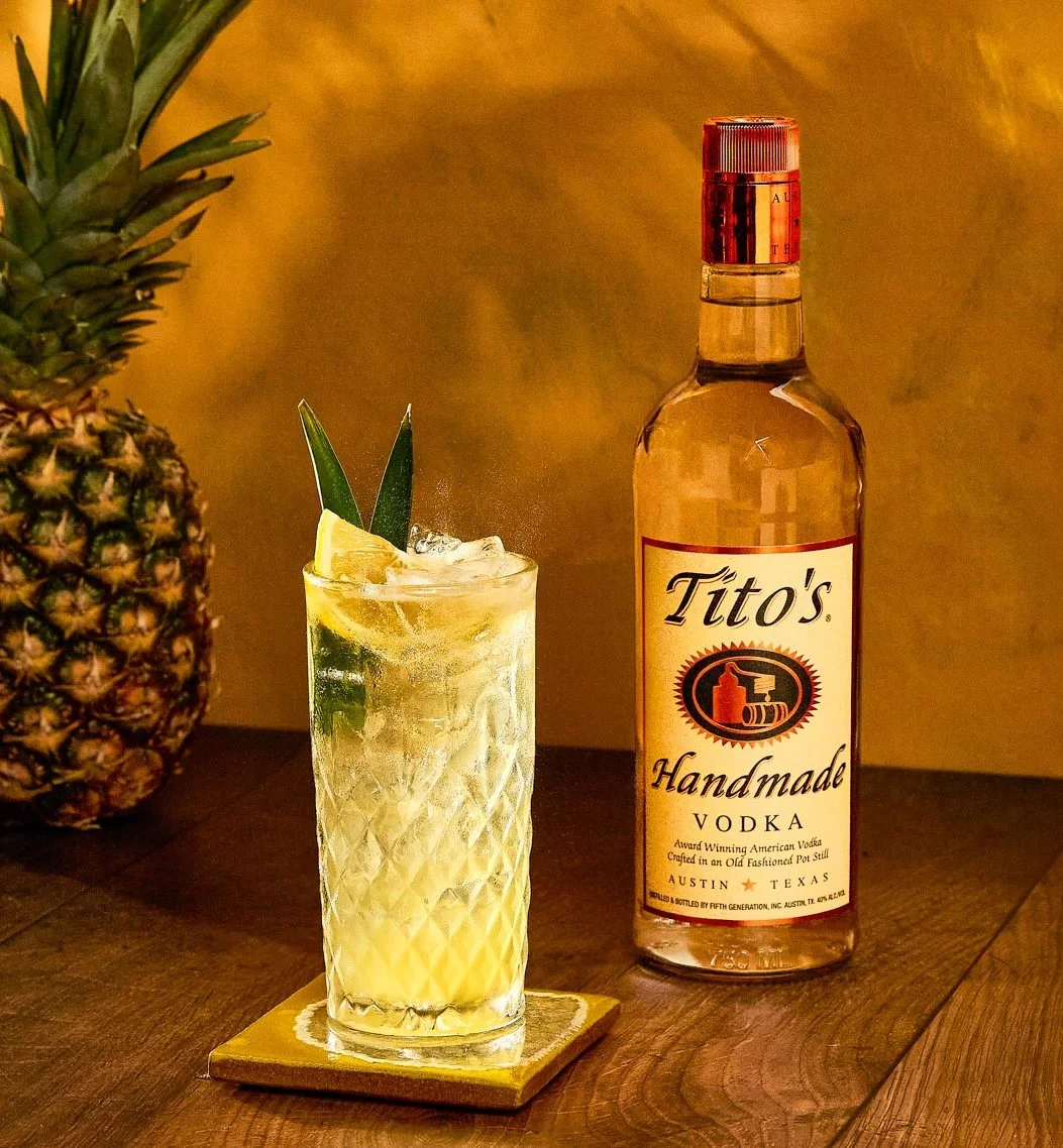 A cocktail glass with pineapple leaves and a lemon wedge, filled with a yellow drink and ice, placed on a coaster next to a bottle of Tito's Handmade Vodka, with a pineapple in the background.