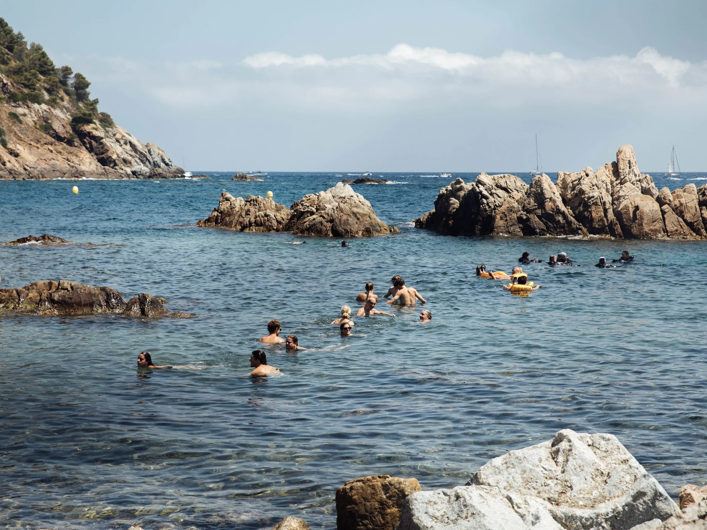 People swimming and relaxing in rocky ocean waters near a coastline with a steep hill covered in trees, and boats on the horizon under a partly cloudy sky.