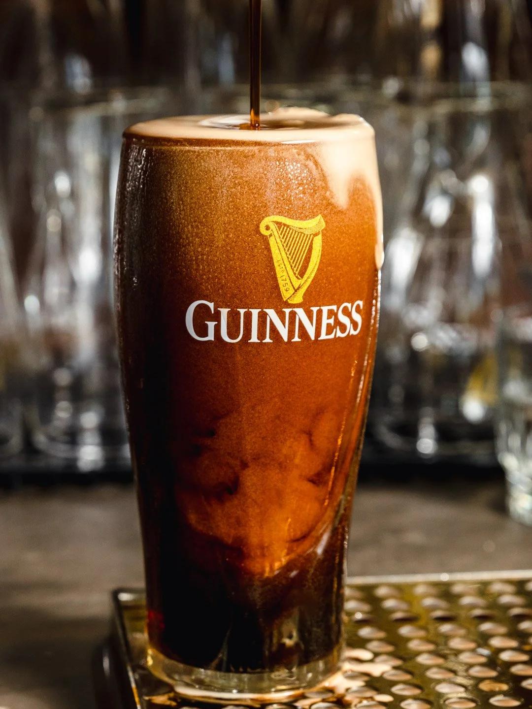 A pint of Guinness beer being poured with foam on top, on a bar counter, with a blurred background of glassware.