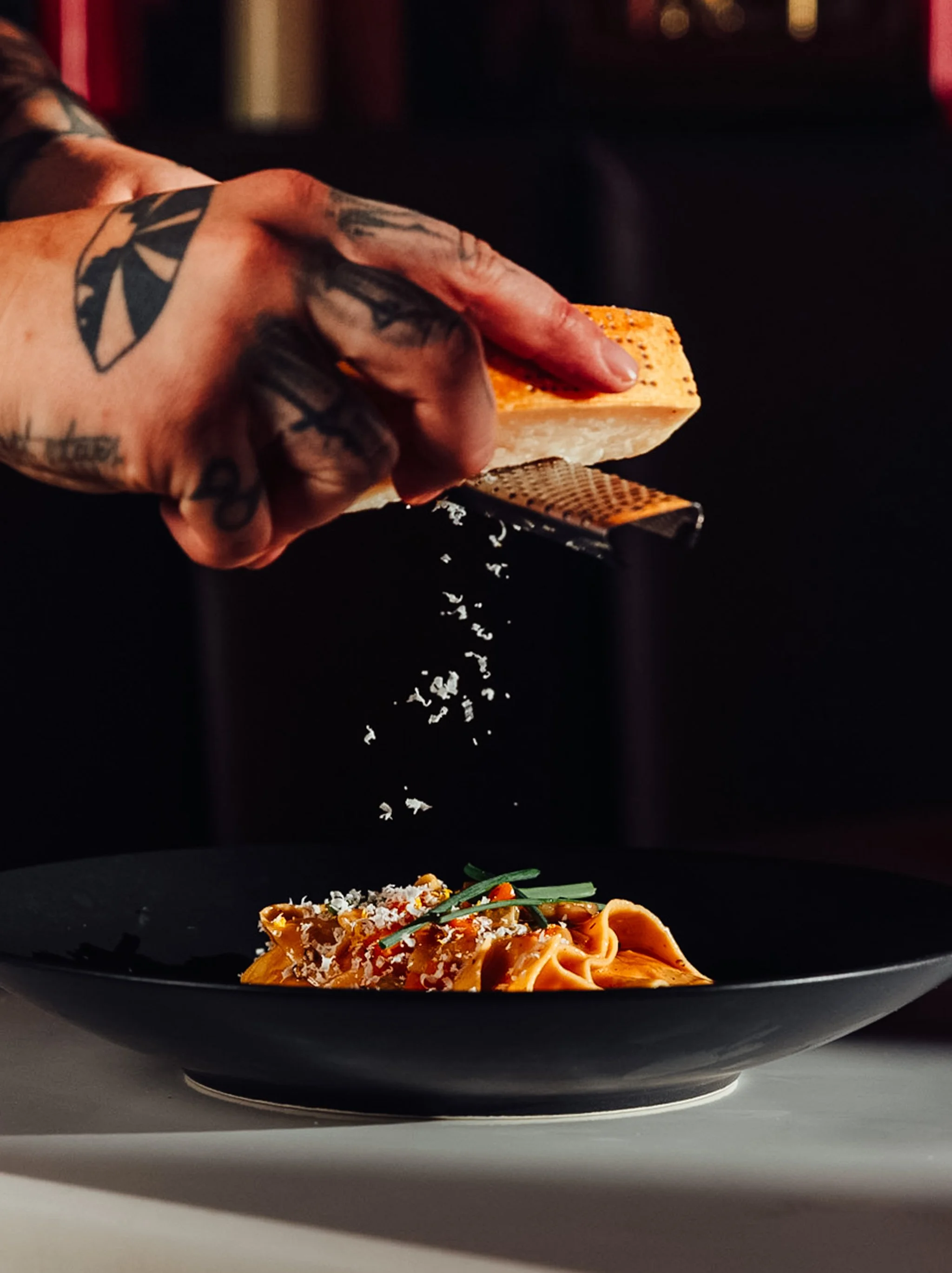 Person grating cheese over a plate of pasta in a restaurant setting.