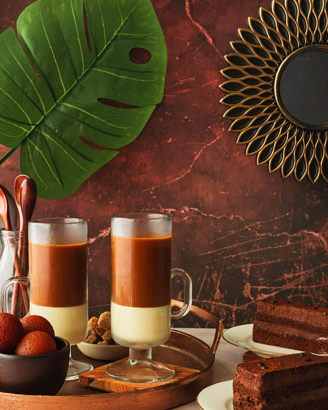 Two glass mugs filled with layered coffee and milk, a bowl of strawberries, chocolate brownies on white plates, and wooden utensils on a tray against a dark red marbled wall with large green leaves and a decorative gold sunburst mirror.