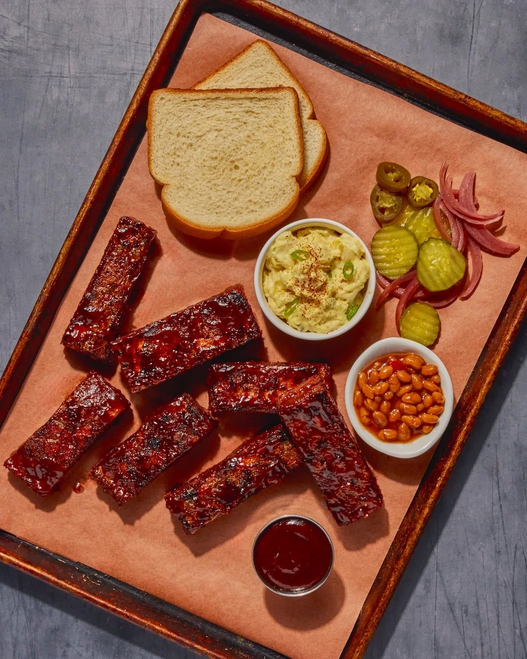 A tray of barbecue ribs, sliced bread, pickles, sliced onions, jalapenos, baked beans, a small bowl of potato salad, and a side of barbecue sauce.