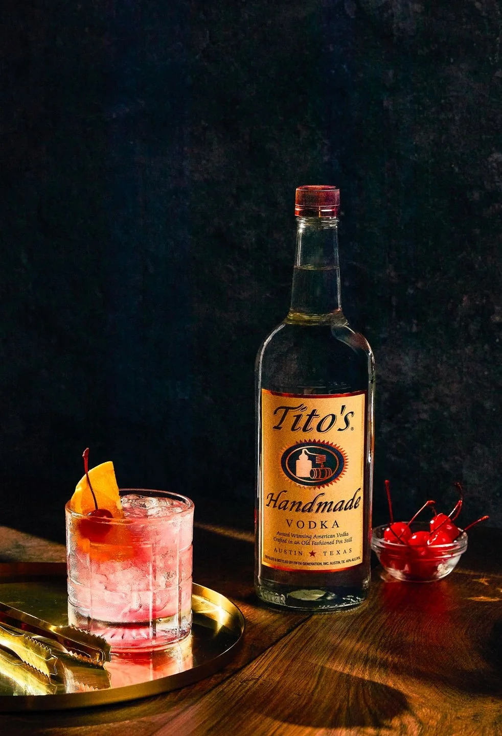 A bottle of Tito's Handmade Vodka, a glass with a pink cocktail garnish with a cherry and orange slice, a small bowl of cherries, and cocktail tongs on a wooden table against a dark background.