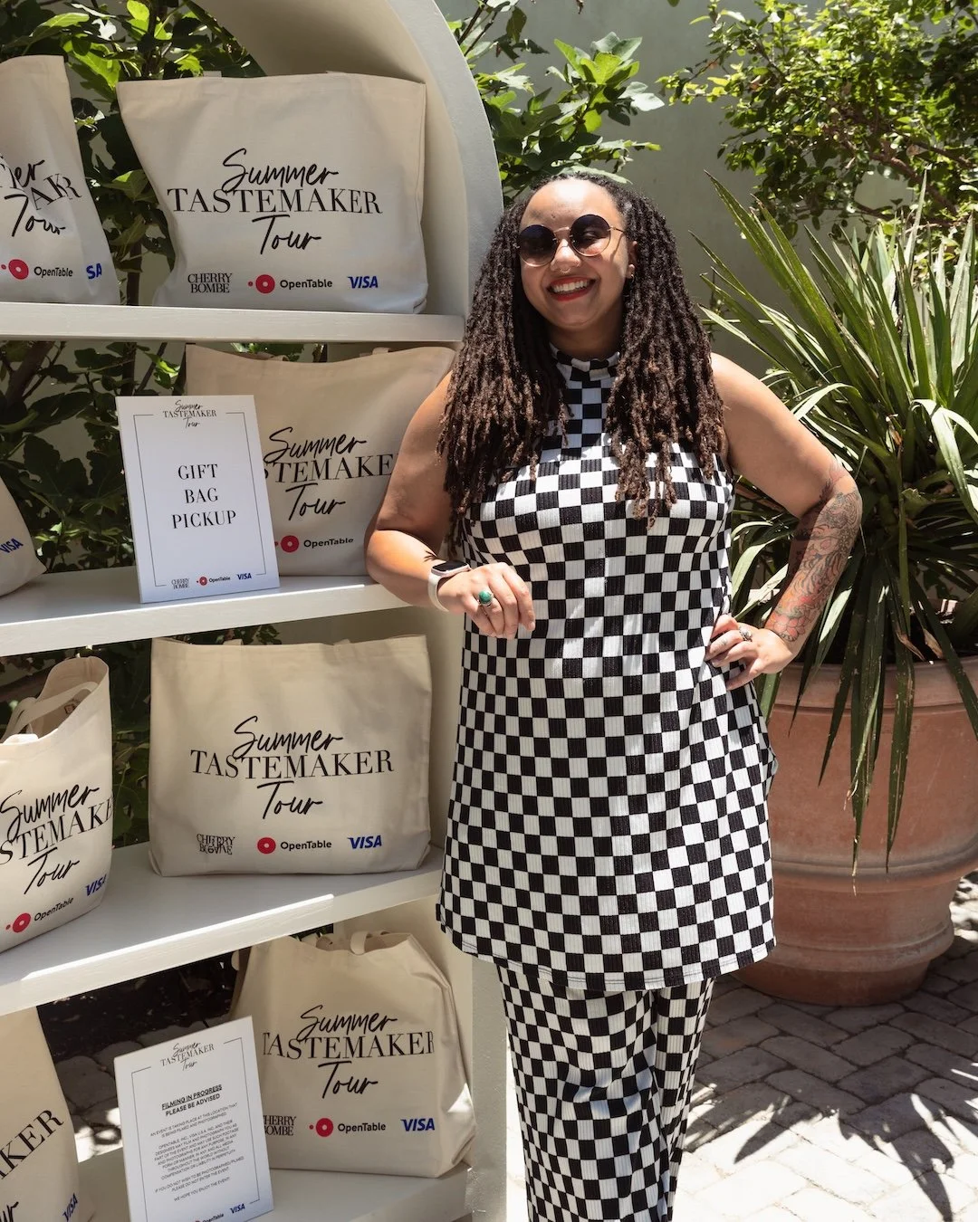 A woman wearing black and white checkered clothing standing outdoors next to a display of tote bags that have the words 'Summer Tastemaker Tour' printed on them. She is smiling, wearing sunglasses and has tattoos on her right arm.