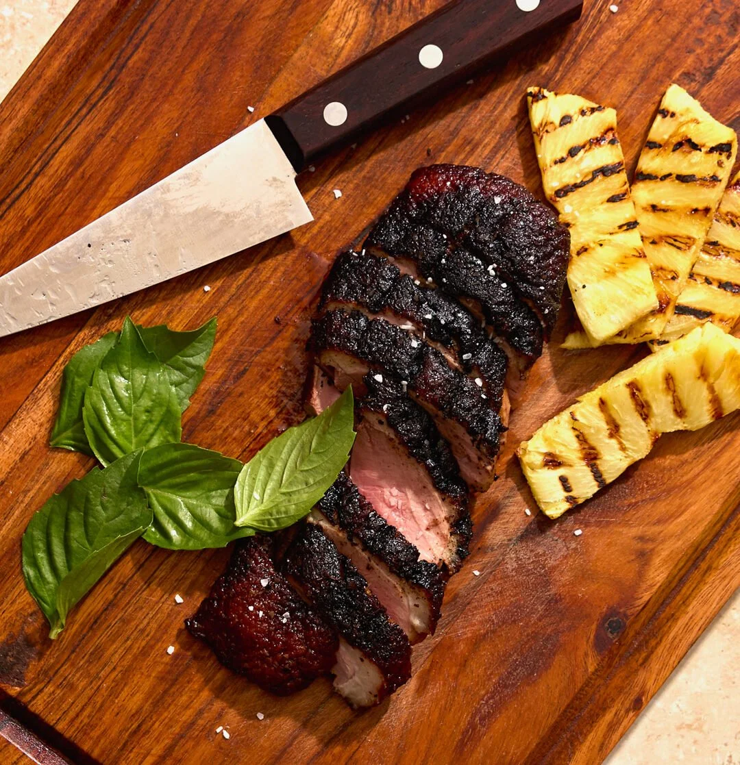 A wooden cutting board with grilled pineapple slices, a sliced seared steak with a dark crust, fresh basil leaves, and a knife.