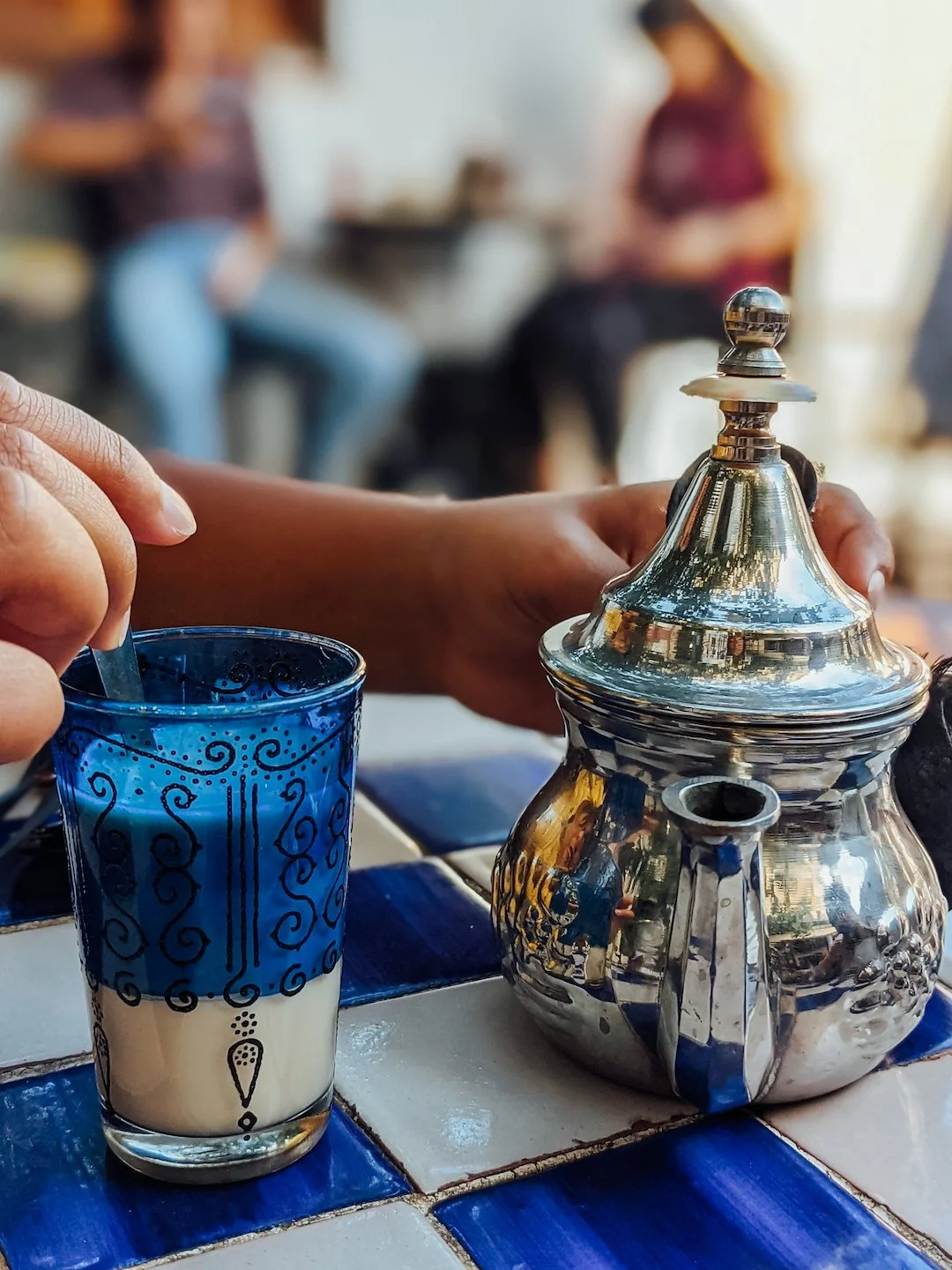 A hand holding a silver teapot, with a blue and white ceramic cup nearby on a tiled table. The background is blurred with people sitting and standing.