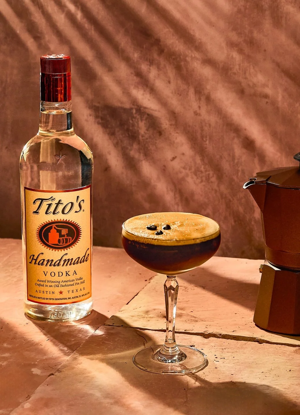 A bottle of Tito's handmade vodka, a cocktail with dark liquid and coffee beans on top in a cocktail glass, and a moka pot on a pink marble surface against a textured pink background.