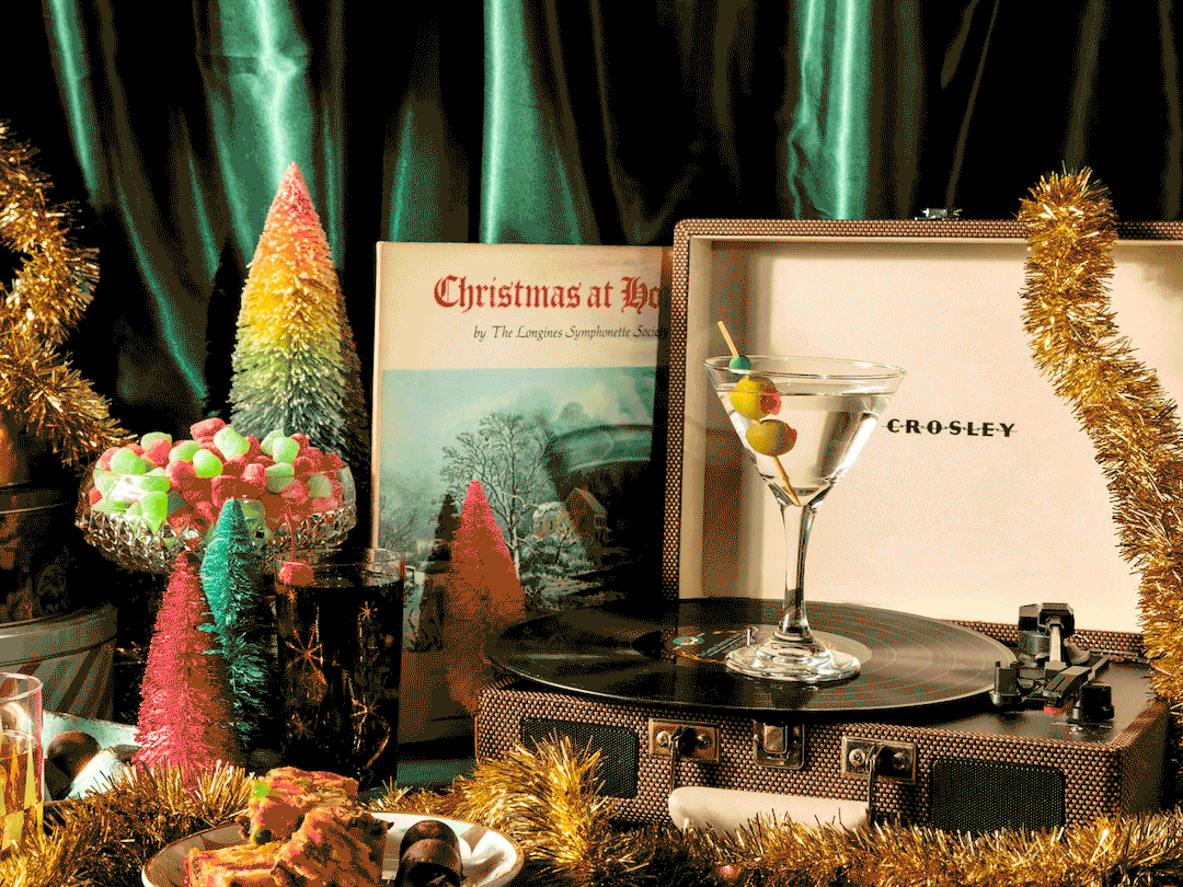 A vintage record player with a glass of martini on top, surrounded by Christmas-themed decorations including small decorated trees, tinsel, candies, and a Christmas album cover in the background.