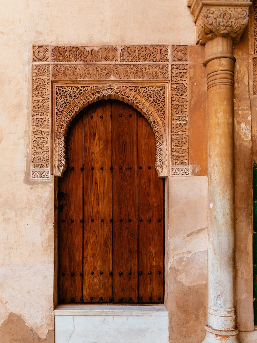 Wooden arched door set in a stone wall with intricate carved stone borders and a decorative column.