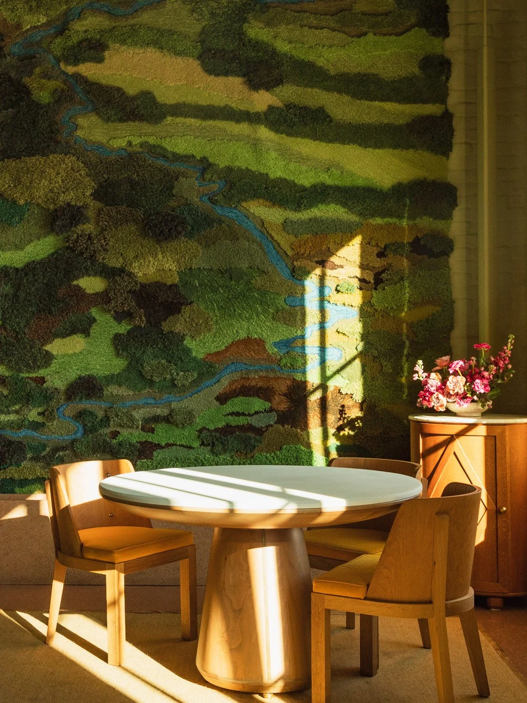 A round wooden table with three wooden chairs is placed in front of a green mountain landscape tapestry on the wall. Sunlight casts shadows over the table, and a wooden cabinet with a flower arrangement in a pink and white vase is set against the wal
