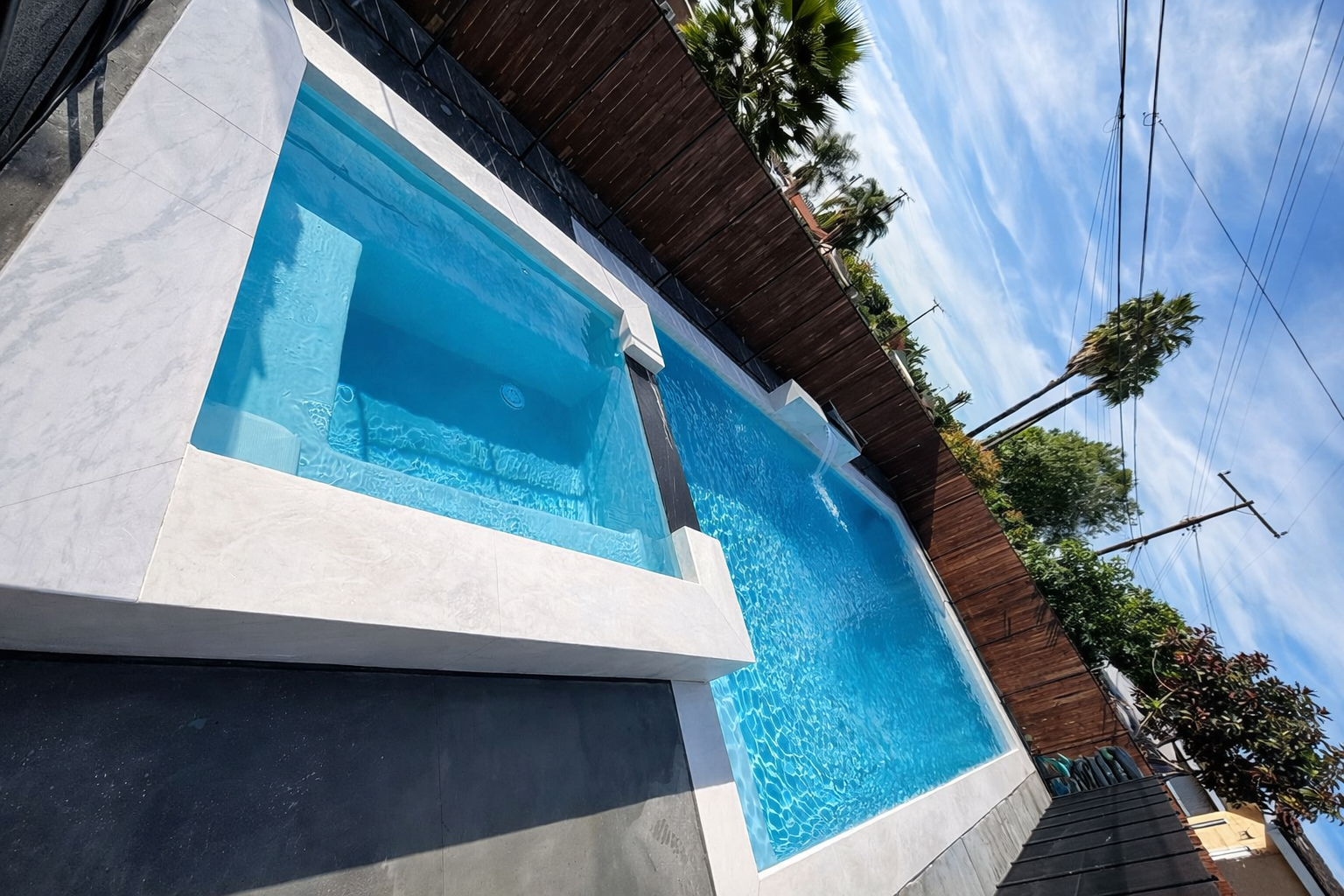 A modern rectangular swimming pool with a spa area, surrounded by a dark deck and a wooden fence, with palm trees and power lines in the background under a blue sky with some clouds.