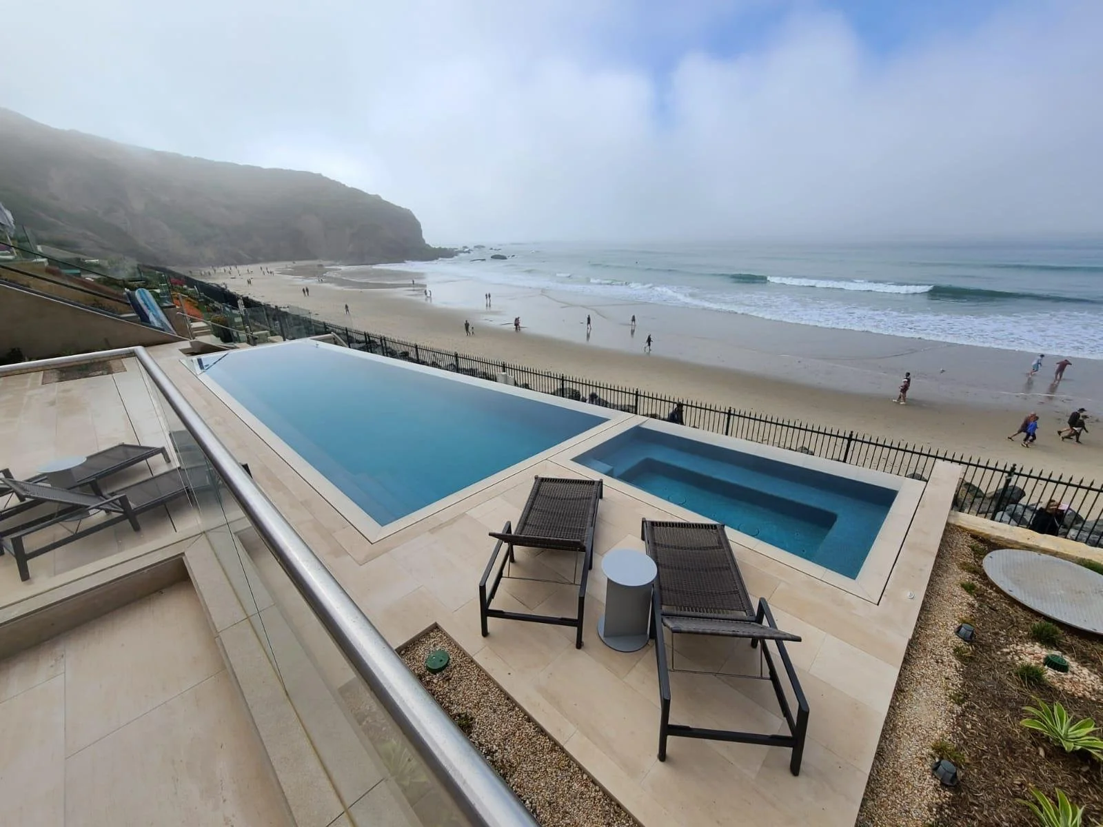 View of a beach with an outdoor pool and hot tub on a terrace, overlooking the ocean with waves and a foggy sky, several people walking along the shore.