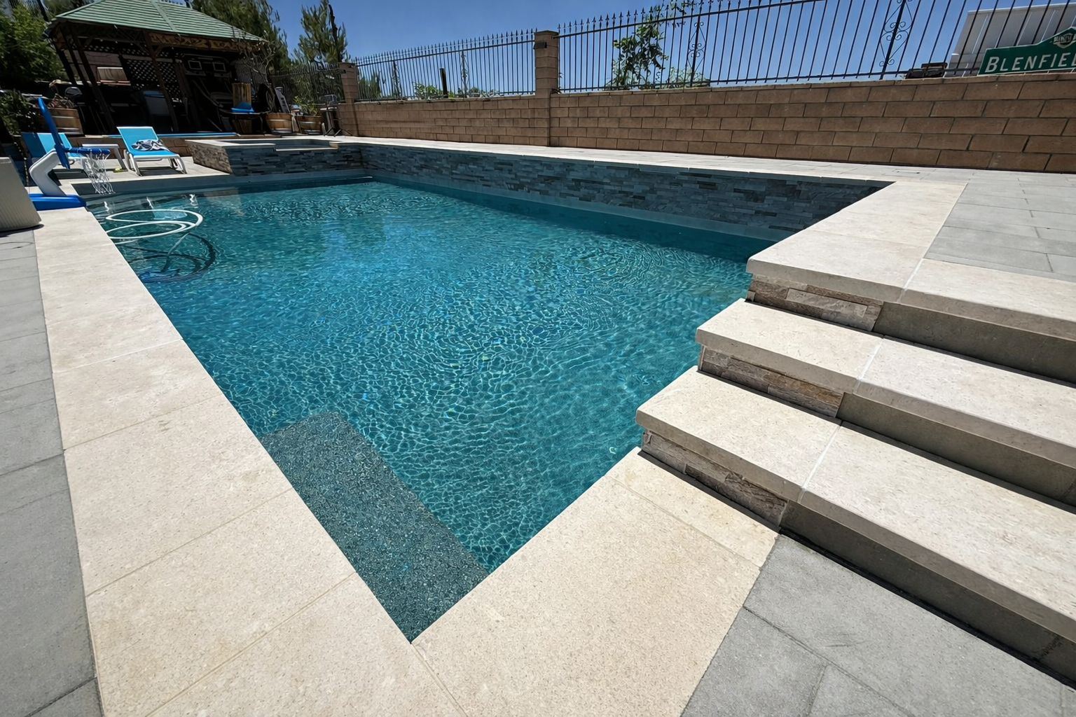 A backyard swimming pool with clear blue water, surrounded by beige and gray tiled deck, and a brick wall with a fence above. There are stairs leading into the pool and poolside chairs in the background.
