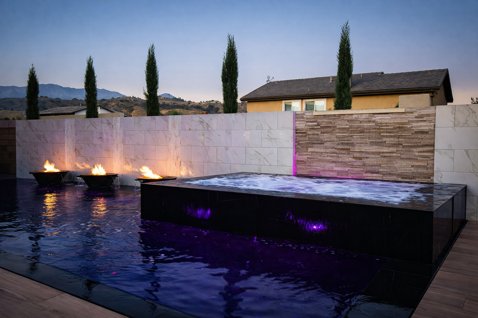 Outdoor modern pool with water features and fire bowls, surrounded by a stone wall with a house and trees in the background at dusk.